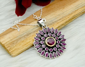 May include: A silver pendant necklace featuring a large, circular pendant with a central, round, deep red gemstone surrounded by smaller, oval-shaped, deep red gemstones. The pendant has a gold-colored border and is on a delicate silver chain.