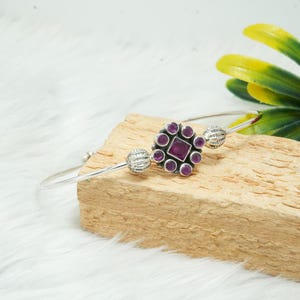 Puede incluir: Un brazalete de plata con un centro de piedra preciosa púrpura rodeado de piedras preciosas púrpuras más pequeñas. El brazalete tiene dos acentos de bola de plata.