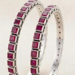 May include: Two circular silver bangles, each featuring a continuous row of square-cut, deep red rubies set in a channel. The metal band has a polished finish, and the stones are arranged in a uniform pattern around the entire circumference.