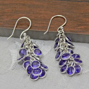 May include: A pair of silver-coloured drop earrings. Each earring has a cluster of small, oval, deep purple gemstones. The gemstones hang from a silver hook, creating a cascading effect. The earrings are displayed on a light grey fabric.