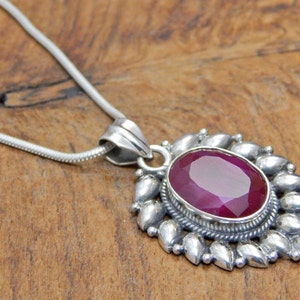 Puede incluir: Un collar con un colgante de plata con una gran piedra de rubí ovalada engastada en un bisel de plata. El colgante está rodeado por un diseño decorativo de plata de pequeñas hojas puntiagudas.