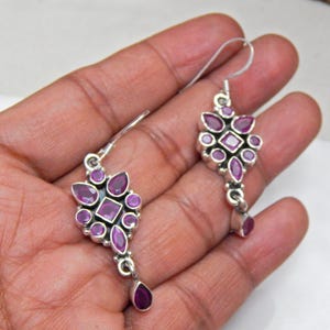 Może przedstawiać: Para srebrnych kolczyków wiszących z gromadą fioletowych kamieni szlachetnych w kwiatowym wzorze. Kolczyki mają kamień szlachetny w kształcie kropli u dołu.