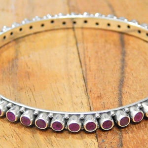 May include: A circular silver bangle bracelet featuring a continuous row of small, round, deep pink cabochon stones set in individual metal bezels. The silver band has a polished finish and a series of small decorative beads between each stone setting.