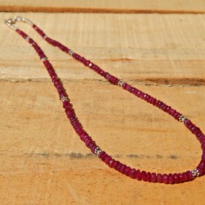 Op de afbeelding: Een rode robijn edelsteen ketting met zilveren kralen. De ketting is gemaakt van kleine, gefacetteerde robijn kralen en zilveren spacer kralen. De ketting ligt op een houten oppervlak.