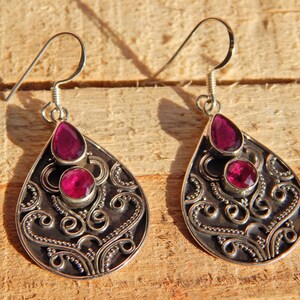 May include: A pair of silver earrings with a teardrop-shaped design. Each earring features two pink gemstones set in the centre of the teardrop.