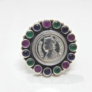 Puede incluir: Un anillo de plata con un diseño de moneda redonda que presenta el perfil de una mujer. La moneda está rodeada por un círculo de piedras preciosas de colores, incluyendo rosa, verde y azul.