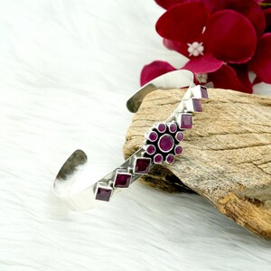 Peut inclure: Un bracelet manchette en argent orné d'un motif floral et de pierres précieuses rubis taillées en carré. Le bracelet présente un groupe central de rubis ronds entourés de rubis plus petits. Le bracelet est présenté sur un morceau de bois avec un fond blanc et une fleur rouge.