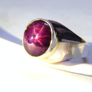 Op de afbeelding: Een zilveren ring met een grote, ovale, rode robijn edelsteen. De robijn heeft een stervormig patroon in het midden.