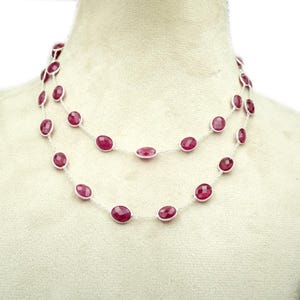 Op de afbeelding: Een ketting met twee strengen met ovale, dieprode edelstenen gezet in een zilveren ketting. De edelstenen zijn gelijkmatig verdeeld, waardoor een gelaagd effect ontstaat. De ketting wordt op een mannequin getoond.