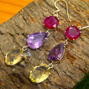 Puede incluir: Un par de pendientes de plata con tres piedras preciosas. La piedra preciosa superior es un rubí rojo redondo. La piedra preciosa del medio es una amatista púrpura en forma de pera. La piedra preciosa inferior es una citrina amarilla ovalada.