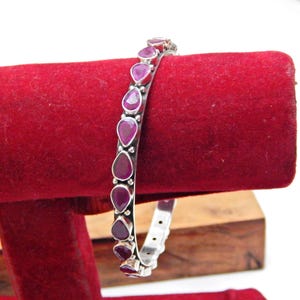 Puede incluir: Un brazalete de plata con piedras preciosas rojas en forma de lágrima. Las piedras están dispuestas en una fila a lo largo de la parte superior del brazalete. El brazalete se muestra sobre una superficie de terciopelo rojo.