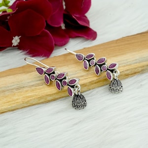 Peut inclure: Boucles d'oreilles pendantes argentées avec un motif floral et de petits breloques en forme de cloche. Les boucles d'oreilles présentent des pierres en forme de feuille rose foncé. Les boucles d'oreilles sont présentées sur une surface en bois avec une fleur bordeaux en arrière-plan.