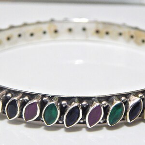 Könnte beinhalten: Ein silbernes Armreif-Armband mit einem dekorativen Design aus abwechselnd grünen, violetten und blauen Steinen.