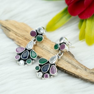 May include: A pair of silver-toned drop earrings with a cluster of colorful gemstones. The earrings feature a mix of round and teardrop-shaped stones in shades of green, purple, blue, and clear, set against a silver metal. The earrings are displayed on a wooden surface.