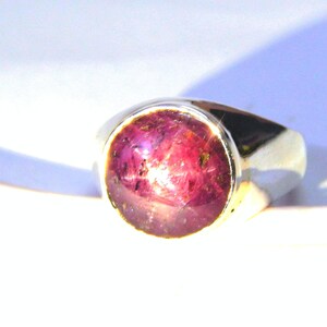 Może przedstawiać: Srebrny pierścionek z okrągłym, różowym rubinem. Rubin ma wzór w kształcie gwiazdy.