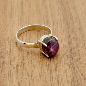 Puede incluir: Un anillo de plata con una gema grande, ovalada y de color rojo oscuro con un patrón de estrella. El anillo tiene una banda lisa y pulida, y la gema está sujeta por cuatro garras. El anillo está sobre una superficie de madera.