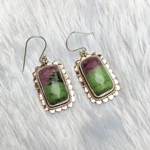 May include: A pair of drop earrings featuring rectangular ruby zoisite gemstones with distinct green and magenta-red coloration. Each stone is set in a scalloped silver frame with a twisted rope border and attached to a simple silver hook wire.