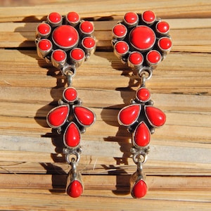 May include: A pair of dangle earrings with red coral stones. The earrings feature a cluster of round stones at the top and a teardrop-shaped cluster of stones hanging below.