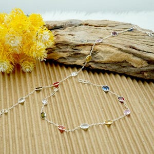 Op de afbeelding: Een zilveren enkelketting met ovale edelstenen in verschillende kleuren, waaronder rood, oranje, geel, groen, blauw en roze.