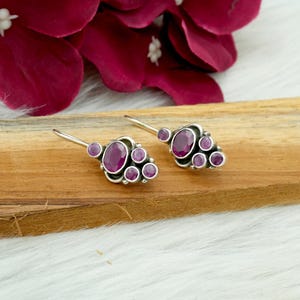Puede incluir: Pendientes de plata con un grupo de gemas ovaladas y redondas de color rojo intenso. Los pendientes se exhiben sobre una superficie de madera, con un fondo de flores de color rosa oscuro.