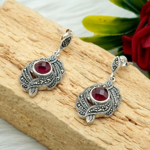 May include: A pair of silver-toned drop earrings with a central, round, deep red gemstone. The earrings have an ornate, leaf-like design with small, sparkling accents. The earrings are displayed on a wooden surface.