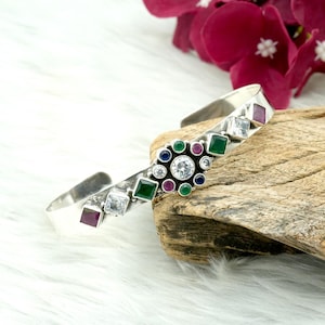 May include: A silver cuff bracelet with a floral design. The bracelet features a central cluster of round and square gemstones in various colours, including clear, green, red, and blue. The bracelet is set on a wooden surface.