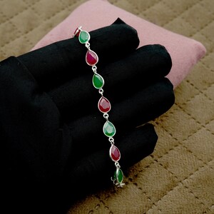 Puede incluir: Una pulsera de plata con piedras preciosas en forma de lágrima alternando rojo y verde. La pulsera se muestra sobre un fondo negro, resaltando los colores vibrantes de las piedras. Es una pieza de joyería.