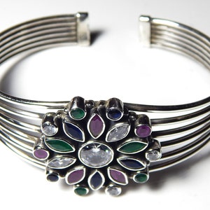 Op de afbeelding: Zilveren armband met een veelkleurig bloemenmotief. Het ontwerp bevat groene, blauwe, roze en witte edelstenen.