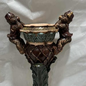 May include: Ornate decorative pedestal with a detailed design. The pedestal features a brown, green, and gold color scheme, with monkey figures supporting the top bowl. The base has a leaf design.