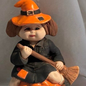 May include: A ceramic figurine of a witch sitting on a pumpkin. The witch is wearing an orange hat with a black band and a black dress. She is holding a broom in her hands. The pumpkin is orange and has a carved jack-o'-lantern face. The figurine is lit from within.