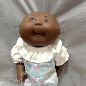 May include: A Cabbage Patch Kid doll with brown skin, brown eyes, and a white ruffled shirt. The doll is wearing blue dungarees with a pink and white floral pattern.