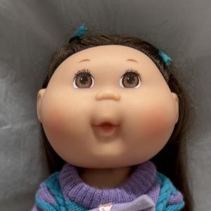 Könnte beinhalten: Eine Cabbage Patch Kid Puppe mit braunen Haaren, die einen lila-blauen gestreiften Pullover mit einer rosa Schleife trägt. Die Puppe trägt weiße Stiefel.