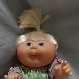 May include: A Cabbage Patch Kid doll with blonde hair in a ponytail, wearing a green and pink floral jumpsuit with a pink and white striped sash.