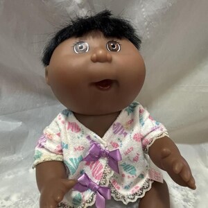 Könnte beinhalten: Eine Cabbage Patch Kid Puppe mit brauner Haut, schwarzen Haaren und braunen Augen. Die Puppe trägt ein weißes und rosa gemustertes Schlafkleid mit lila Schleifen.