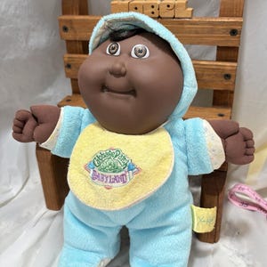 Peut inclure: Une poupée Cabbage Patch Kids Babyland à la peau brune, aux cheveux bruns et aux yeux bruns. La poupée porte une combinaison bleue à capuche avec une bavette jaune sur laquelle est inscrit "Cabbage Patch Kids Babyland".