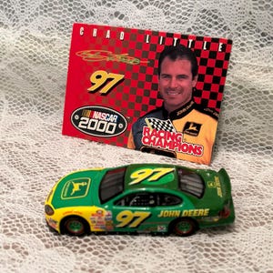 May include: A green and yellow toy race car with the number 97 and John Deere logos. A red card with the text "CHAD LITTLE" and a photo of a man in a racing uniform is behind the car. The card also says "NASCAR 2000".