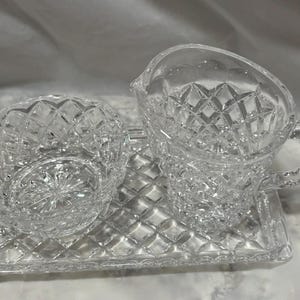 May include: A clear crystal creamer and sugar bowl set with a diamond pattern, resting on a matching rectangular tray. The creamer has a handle and spout. The set is designed for serving.