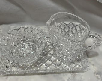 May include: A clear crystal creamer and sugar bowl set with a diamond pattern, resting on a matching rectangular tray. The creamer has a handle and spout. The set is designed for serving.
