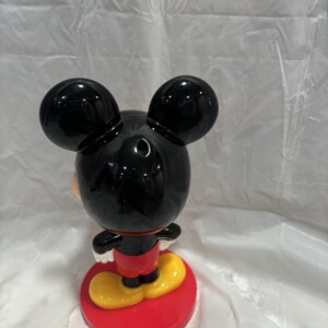 K&ouml;nnte beinhalten: Eine Mickey Mouse-Figur, von hinten gesehen, mit gl&auml;nzendem schwarzem Kopf und Ohren. Die Figur tr&auml;gt ein rotes Hemd, gelbe Schuhe und steht auf einem roten kreisf&ouml;rmigen Sockel. Die Figur ist ein Sammlerst&uuml;ck.
