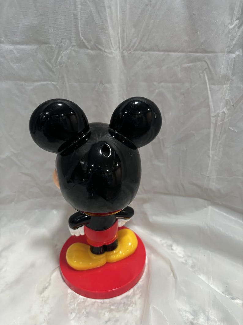 K&ouml;nnte beinhalten: Eine Mickey Mouse-Figur, von hinten gesehen, mit gl&auml;nzendem schwarzem Kopf und Ohren. Die Figur tr&auml;gt ein rotes Hemd, gelbe Schuhe und steht auf einem roten kreisf&ouml;rmigen Sockel. Die Figur ist ein Sammlerst&uuml;ck.