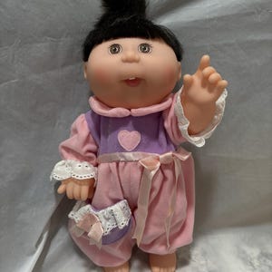 May include: A Cabbage Patch Kid doll with black hair, brown eyes, and a pink and purple outfit. The doll is wearing a pink and white lace trim on the sleeves and a pink ribbon around the waist.