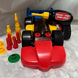 May include: A colorful toy race car set with a black power drill. The car is red, yellow, and blue with black wheels. The set includes yellow and red screws, yellow nuts, and a green disc. The power drill is black with the text "Construct Power Power Drill".
