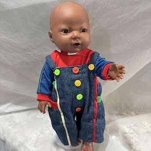 May include: A brown baby doll wearing a blue and red long-sleeved shirt with a denim overall with colorful buttons.