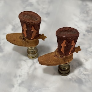 Peut inclure: Deux embouts de lampe en forme de botte de cowboy marron avec des accents orange. Les embouts de lampe sont en céramique et ont un aspect rustique.