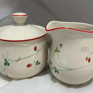 Op de afbeelding: Crèmekleurige keramische suikerpot met deksel en melkkan. Beide stukken zijn versierd met een aardbeienrankenontwerp, met rode aardbeien, witte bloemen en groene bladeren. De randen zijn rood afgewerkt.