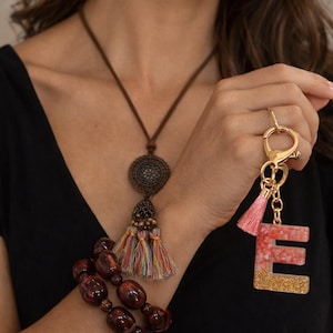May include: A woman is wearing a brown necklace with a circular pendant and colorful tassels. A red beaded bracelet and a gold keychain with a pink and gold letter "E" are also visible. The woman is wearing a black top.