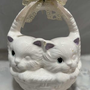 May include: White ceramic basket shaped like two cats facing each other. The basket has a handle and is decorated with a pink rose, green leaves, and lace ribbon. The cats have blue eyes and purple accents.