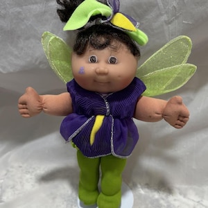 May include: A Cabbage Patch Kid doll dressed as a fairy with green wings, a purple dress, and green leggings. The doll has brown hair, brown eyes, and a painted-on ladybug on her cheek.