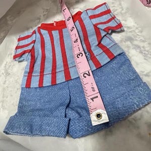 May include: A blue and red striped shirt with a blue denim shorts outfit for a doll. The outfit is laid flat on a white surface with a pink measuring tape.