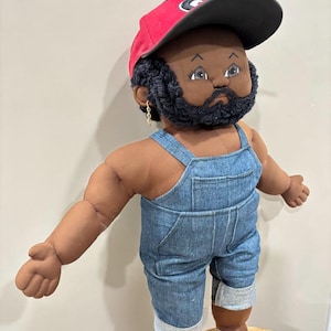 Puede incluir: Muñeca hecha a mano con piel oscura, que lleva una gorra de béisbol roja con visera negra y un peto vaquero. La muñeca tiene barba y bigote negros, y pendientes dorados. La muñeca está sobre una superficie de madera.